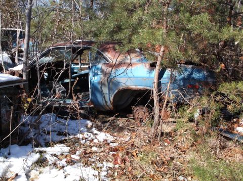 1974 Dodge Challenger - A rusty, weathered blue classic car sits abandoned in a wooded area, partially obscured by trees and pine needles. Snow and fallen leaves c…
