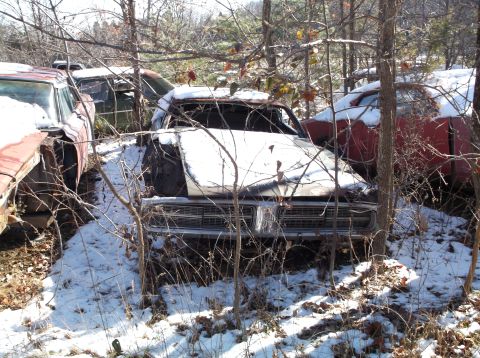 1974 Dodge Challenger - Several old cars are parked in a wooded area with snow on the ground. The front of a white car with a chrome grille is visible, along with…