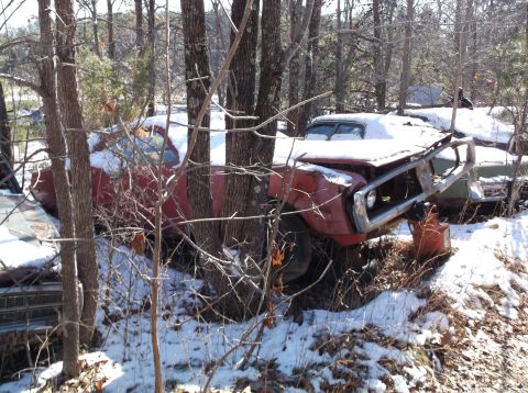 1974 Dodge Challenger - Two snow-covered vintage cars, one red and one green, are parked amongst trees in a wooded area. The red car in the foreground has a damage…