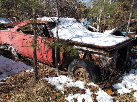 1974 Dodge Challenger - A heavily rusted, red vintage car sits abandoned in a wooded area, partially covered in snow. The vehicle's front end is heavily damaged, w…