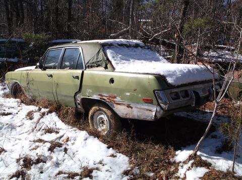 1974 Dodge Challenger - A light green, four-door sedan sits in a snowy, wooded area. The vehicle shows significant rust and damage, particularly around the rear wh…