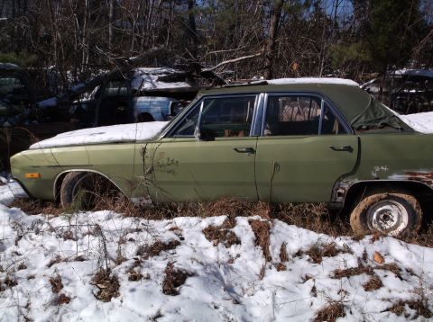 1974 Dodge Challenger - A weathered, light green four-door sedan sits partially covered in snow in a wooded junkyard. The car shows significant rust and damage, pa…