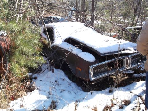 1974 Dodge Challenger - A rusty, snow-covered Dodge Challenger sits abandoned in a wooded area, with its hood open and the interior exposed to the elements. The fr…