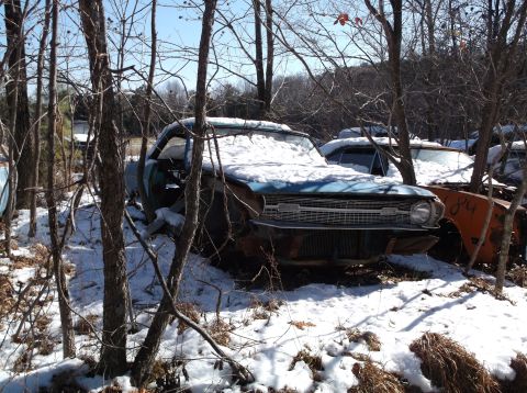 1974 Dodge Challenger - A blue classic car, partially covered in snow, sits in a wooded area with other vehicles in the background. The front grille and headlight…