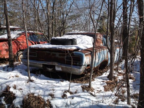 1974 Dodge Challenger - Two heavily rusted, snow-covered vintage cars are parked in a wooded area. The car in the foreground is a blue and rust-colored coupe with…