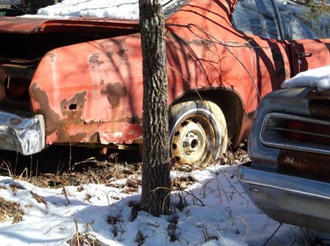 1974 Dodge Challenger - The rear quarter panel and wheel of a rusty red vintage car are visible, partially covered by snow and a tree trunk. Another vintage car's…