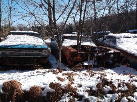 1974 Dodge Challenger - Two abandoned vintage cars, a blue sedan and a rusty orange coupe, are covered in snow in a wooded junkyard setting. The vehicles appear to…