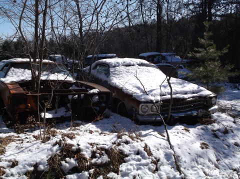 1974 Dodge Challenger - Two rusty, snow-covered classic cars are parked in a wooded area with bare trees. The car in the foreground on the left is missing its hood…