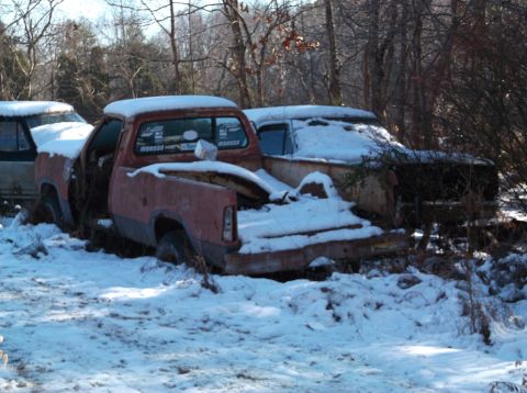 1974 Dodge Challenger - Two old, rusty vehicles sit covered in snow in a wooded area. The vehicle in the foreground is a red pickup truck, and behind it is a white…