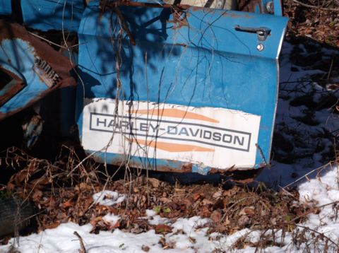1974 Dodge Challenger - A close-up view of a weathered blue vehicle door with a Harley-Davidson logo. The door is rusted and covered in debris, with snow on the gr…