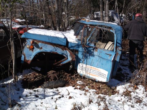 1974 Dodge Challenger - A rusty blue truck cab with a Harley-Davidson decal on the door sits in a snowy junkyard surrounded by trees. A person wearing a red hat an…