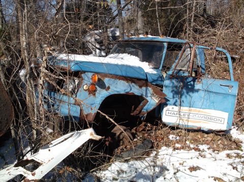 1974 Dodge Challenger - A heavily rusted, blue Dodge pickup truck is overgrown with branches and has snow on its hood. A Harley-Davidson decal is visible on the si…
