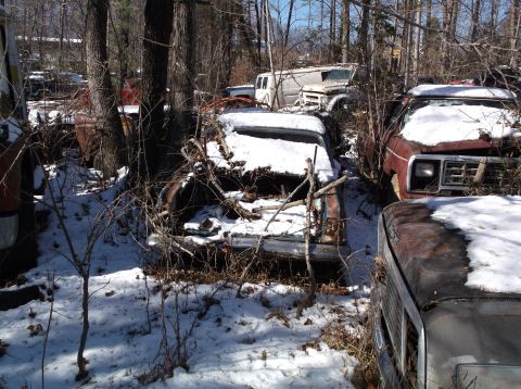 1974 Dodge Challenger - A collection of old, rusted vehicles covered in snow and surrounded by trees. Several cars and a van are visible, some with significant dam…