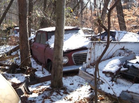 1974 Dodge Challenger - Two old vehicles, a red sedan and a white pickup truck, sit partially covered in snow in a wooded area. The red sedan's front end with its…