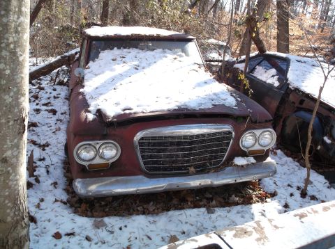 1974 Dodge Challenger - A weathered, red vintage car sits partially covered in snow and fallen leaves in a wooded area. The front of the car features a chrome gril…
