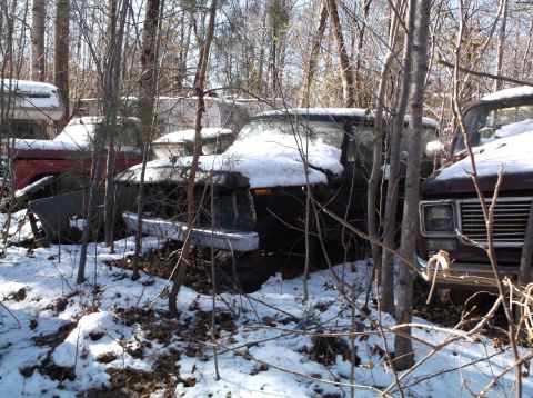 1974 Dodge Challenger - Several old, snow-covered vehicles are parked in a wooded area. The most prominent vehicle in the foreground is a dark-colored pickup truck…