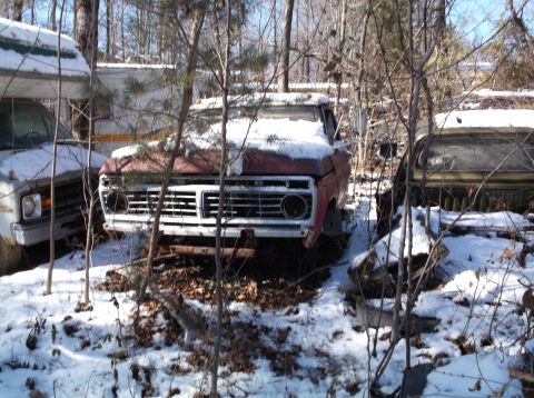 1974 Dodge Challenger - A red pickup truck sits in a snowy junkyard next to a yellow truck. The front of the red truck is visible, showing its grille and headlight…
