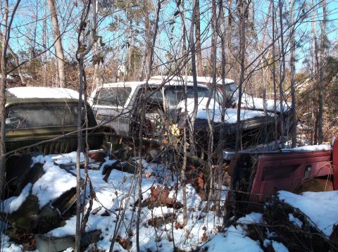 1974 Dodge Challenger - Two old, rusted vehicles are partially covered in snow and surrounded by bare trees. The vehicle on the left appears to be a pickup truck,…
