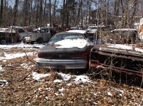 1974 Dodge Challenger - A dark colored sedan sits in a junkyard with snow on its roof and hood, surrounded by trees and other abandoned vehicles.