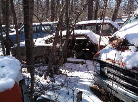 1974 Dodge Challenger - Several old, rusted vehicles are parked in a wooded area covered in snow. One vehicle in the foreground has its hood open and is partially…