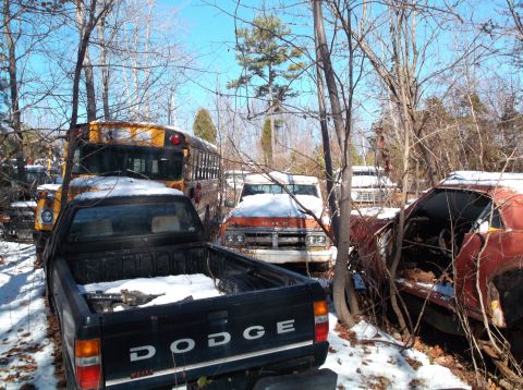 1974 Dodge Challenger - A black Dodge pickup truck is parked in front of a yellow school bus in a snowy junkyard setting. A partially visible red car is to the rig…