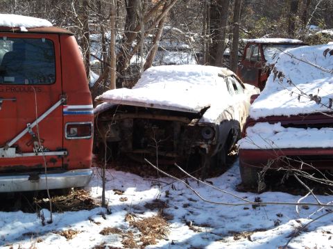 1974 Dodge Challenger - An abandoned orange van is parked next to a snow-covered, rusted-out car in a wooded area. The van has text that reads "G PRODUCTS" on its…