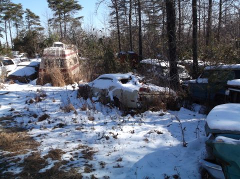 1974 Dodge Challenger - Several old vehicles and a camper sit abandoned in a snowy, wooded junkyard.