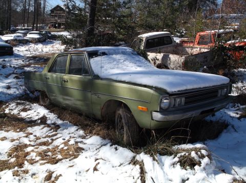 1974 Dodge Challenger - A green sedan sits in a snowy junkyard, surrounded by trees and other abandoned vehicles.