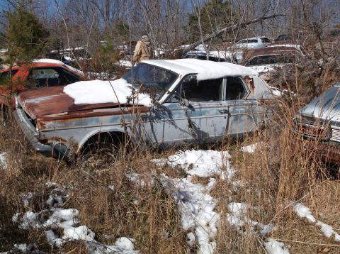 1974 Dodge Challenger - A rusty, light blue sedan sits in a junkyard with snow on the ground and on the car. The car's hood is heavily rusted, and the windows are…