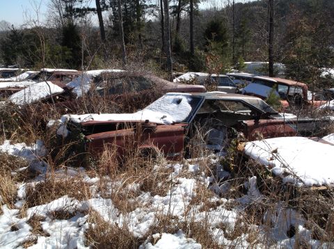 1974 Dodge Challenger - Several old, rusty cars are parked in a wooded area covered in snow. The foreground car is a reddish-brown coupe with its hood and door ope…