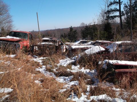 1974 Dodge Challenger - A rusted red 1974 Dodge Challenger sits abandoned in a snowy, overgrown field surrounded by trees. The vehicle is partially covered in snow…