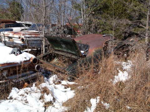 1974 Dodge Challenger - Several rusty, abandoned cars are overgrown with weeds and snow in a junkyard setting. The car in the foreground has its hood open, reveali…