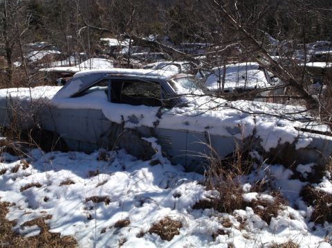 1974 Dodge Challenger - A light blue four-door sedan is partially covered in snow and surrounded by bare trees and brush. The vehicle appears to be in a junkyard o…