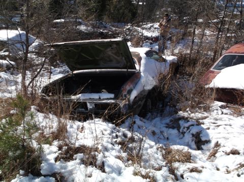 1974 Dodge Challenger - A vintage Dodge Challenger, covered in snow, sits in an outdoor junkyard with its trunk open. A person stands in the background near anothe…