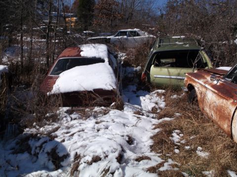 1974 Dodge Challenger - Several old, abandoned cars are covered in snow and overgrown with dry brush in a junkyard setting. The closest vehicle is a rusty brown ca…
