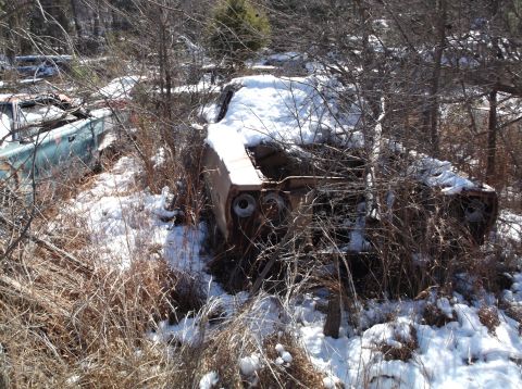 1974 Dodge Challenger - Two abandoned vehicles are partially covered in snow and overgrown with brush. The vehicle in the foreground is heavily rusted and appears…