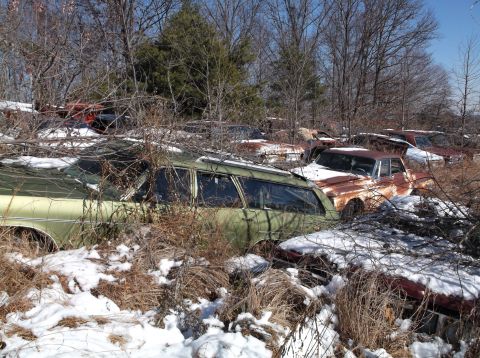 1974 Dodge Challenger - A collection of old, rusted cars are parked in a wooded area covered in snow and dry brush. The foreground features a green station wagon w…