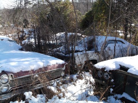 1974 Dodge Challenger - Several abandoned vehicles are covered in snow and overgrown with vegetation. The front of a red car with a chrome grille and dual headligh…