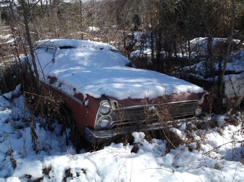1974 Dodge Challenger - A red vintage car, partially covered in snow, sits abandoned in a wooded area with dry brush and trees. The front of the car, including its…