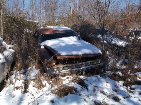 1974 Dodge Challenger - An old, rusty brown car is partially covered in snow and overgrown with brush. The front grille and headlights are visible, as is the hood…