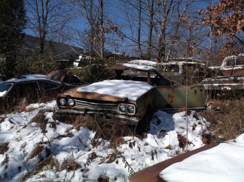 1974 Dodge Challenger - A rusty, abandoned coupe with its hood covered in snow sits in a junkyard surrounded by trees and other derelict vehicles. The car's interi…