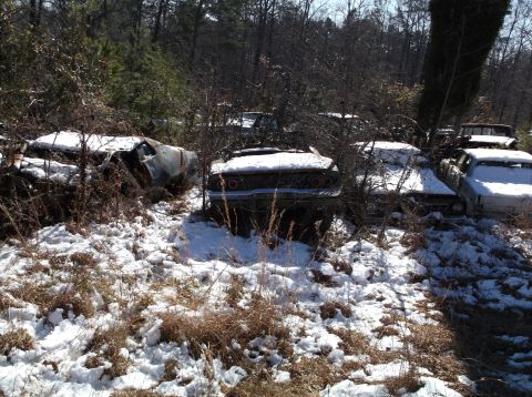 1974 Dodge Challenger - Several old, rusted cars are parked in a wooded area covered in patchy snow. The rear of a dark-colored muscle car with round taillights is…