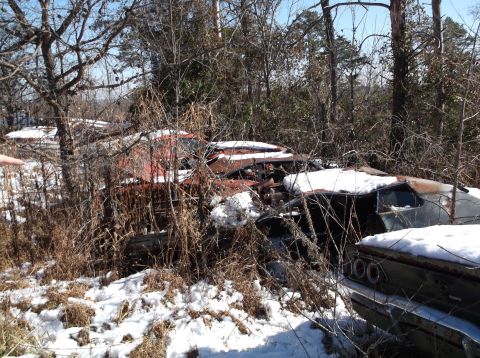 1974 Dodge Challenger - Two rusted vintage cars sit abandoned in a wooded area, covered in snow and overgrown with dry brush. The car in the foreground is a dark c…