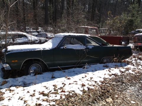 1974 Dodge Challenger - A dark green coupe sits in a junkyard covered in snow, with other vehicles partially visible in the background.