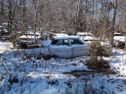 1974 Dodge Challenger - A white sedan sits partially covered in snow in a wooded junkyard. Other vehicles and trees surround the car.