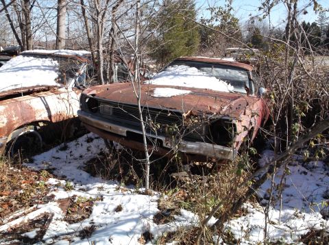 1974 Dodge Challenger - Two rusty, abandoned cars are partially covered in snow and surrounded by bare trees and brush. The car in the foreground is a red coupe wi…