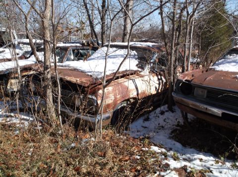 1974 Dodge Challenger - Two rusty, snow-covered classic cars are parked outdoors in a wooded area. The car in the foreground on the right appears to be a 1970s era…