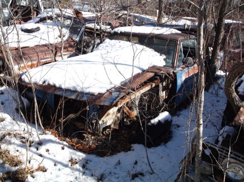 1974 Dodge Challenger - A rusty, snow-covered car sits abandoned in a wooded area, with branches obscuring much of its blue and brown body. The front end is heavil…