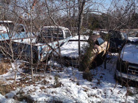 1974 Dodge Challenger - Several rusty, abandoned vehicles are partially covered in snow and surrounded by bare trees. The closest vehicle appears to be a light blu…