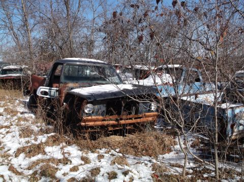 1974 Dodge Challenger - A rusty, dark-colored pickup truck sits in a junkyard with snow on the ground and bare trees surrounding it. Another blue pickup truck is v…